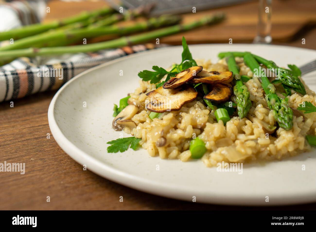 45º vue d'un risotto aux champignons et aux asperges servi sur un plat blanc avec une fourchette, un verre de vin blanc et un râpe et des asperges sur une planche à découper, Banque D'Images
