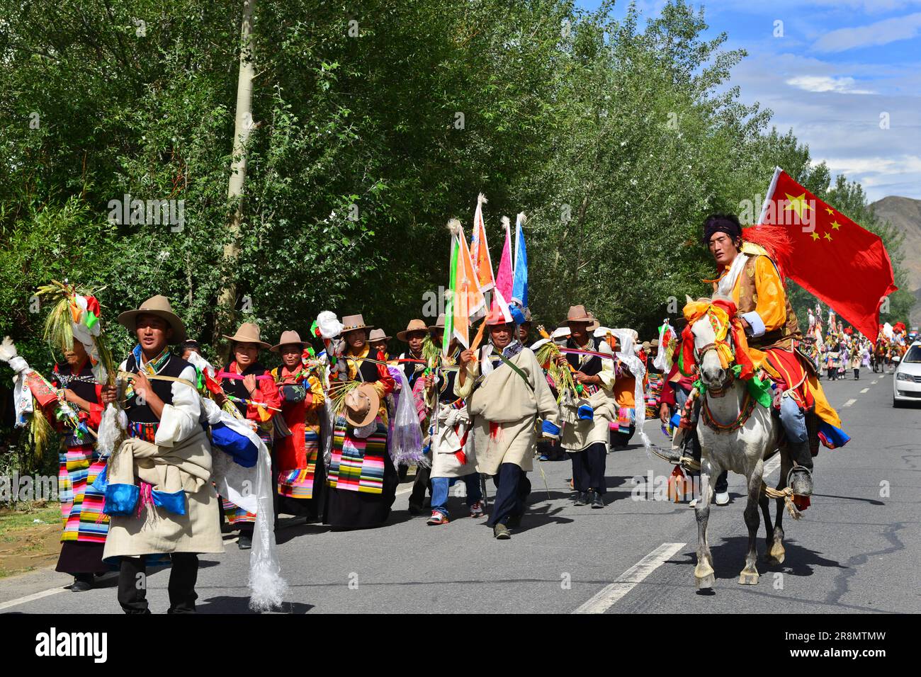 Une procession culturelle dynamique avec des Tibétains habillés traditionnellement marchant le long d'une route, menés par un cavalier portant un drapeau, célébrant Banque D'Images