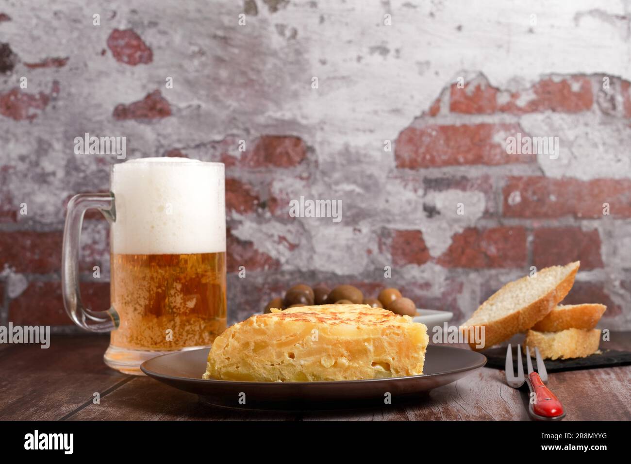 Omelette espagnole typique fraîchement préparée sur une table en bois avec un pichet frais de bière, couverts, pain et olives Banque D'Images