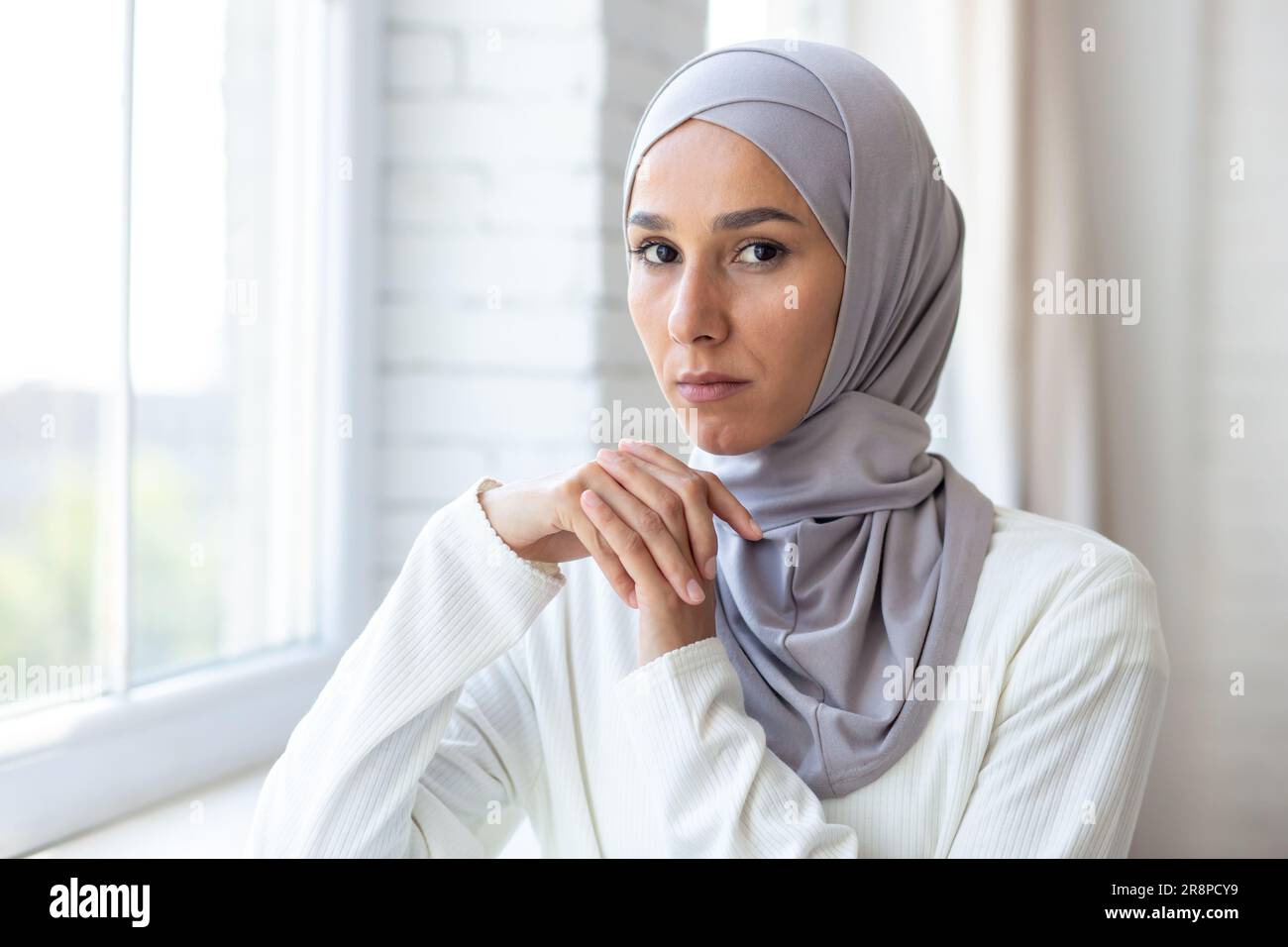 Gros plan. Portrait d'une jeune belle musulmane dans un hijab blanc debout à l'intérieur près de la fenêtre. Il regarde avec confiance et sérieux l'appareil photo. Banque D'Images