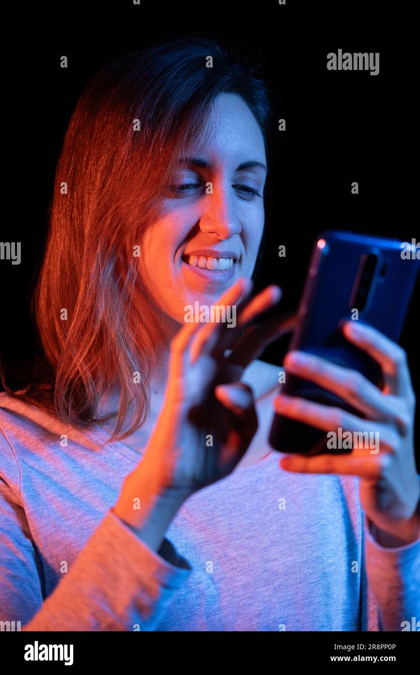 Une jeune fille s'est engarée dans son smartphone, entourée d'un arrière-plan noir mystérieux, illuminée par des lumières bleues et rouges vibrantes pendant qu'elle s'éloigne. Banque D'Images