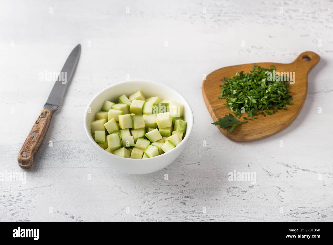 Un bol de courgettes coupées en dés, une planche en bois avec des herbes et un couteau sur fond bleu clair. Cuisine délicieux petit déjeuner maison. Banque D'Images