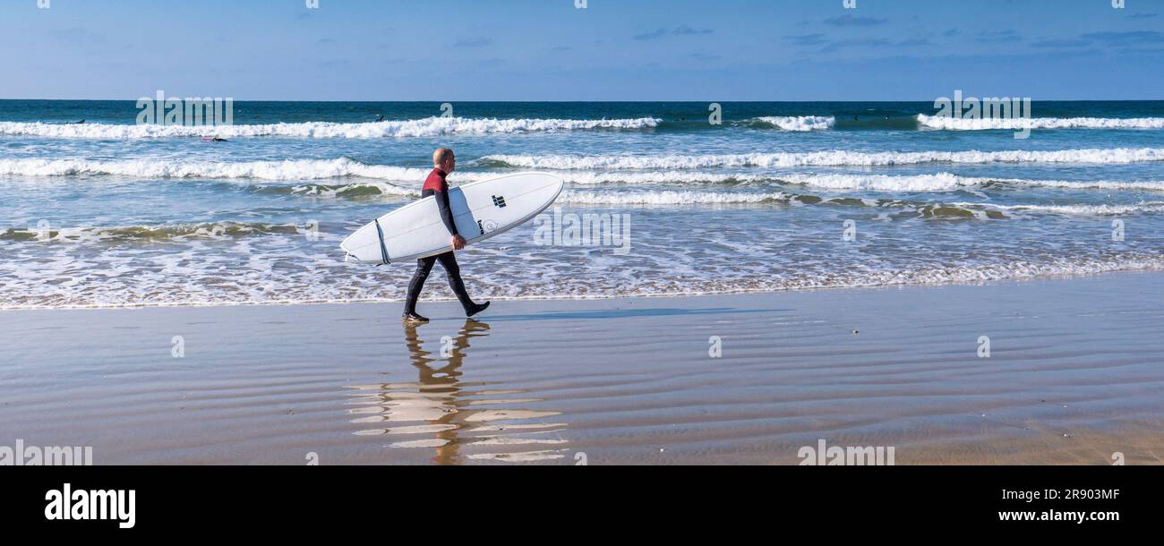Image panoramique d'un surfeur transportant une planche de surf Torq marchant le long du rivage à Fistral à Newquay, en Cornouailles, au Royaume-Uni. Banque D'Images