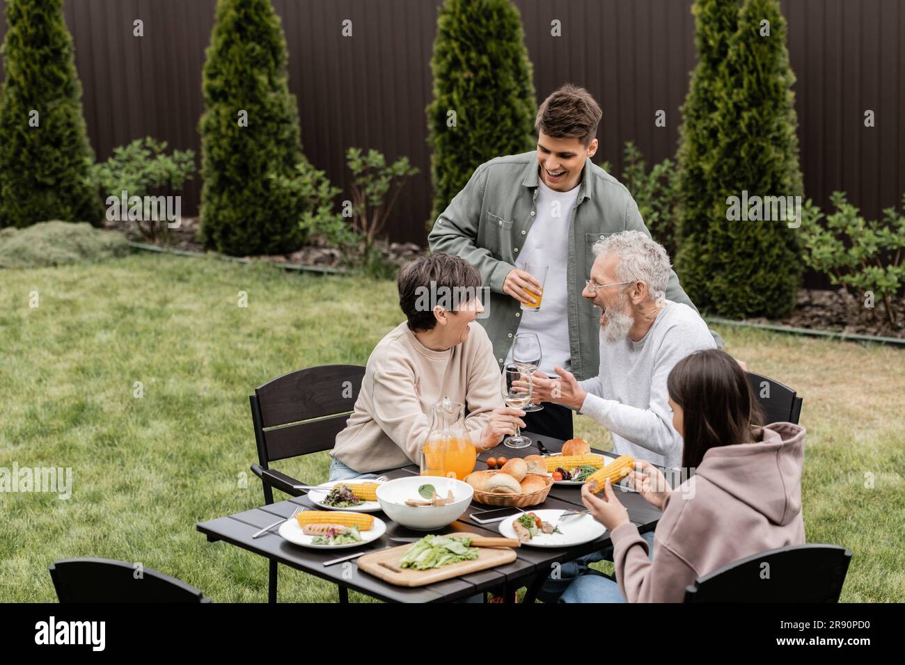 Parents d'âge moyen agités et gaies en train de griller avec des verres à vin près des enfants et de la nourriture d'été pendant la fête de barbecue à l'arrière-cour, chérissant lien familial Banque D'Images