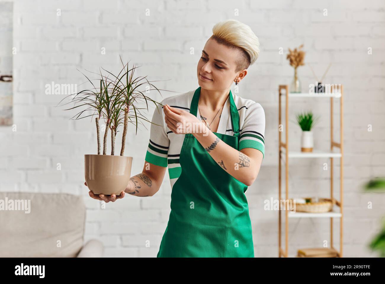 jardinage intérieur, jeune femme positive tatouée dans un tablier vert touchant les feuilles de plantes tropicales dans un appartement moderne, soin des plantes, maison durable Banque D'Images