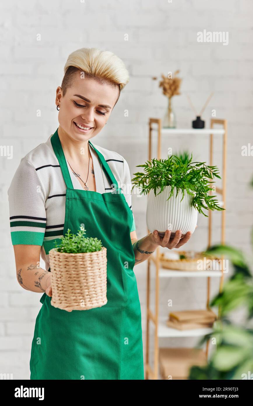 amoureux des plantes, mode de vie éco-conscient, femme tatouée heureuse avec sourire radieux tenant pots de fleurs avec des plantes vertes dans l'appartement moderne, durable Banque D'Images