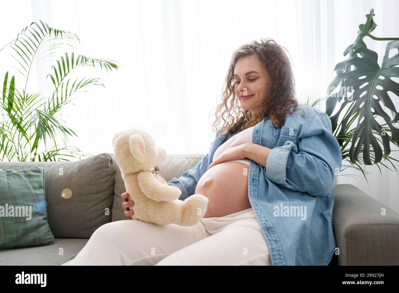 Femme enceinte souriante tenant un ours en peluche assis sur un canapé à la maison Banque D'Images