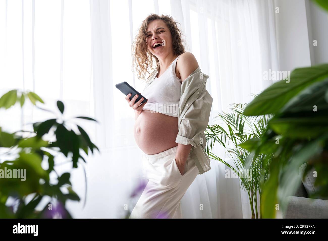 Femme enceinte heureuse avec main dans la poche tenant un smartphone à la maison Banque D'Images