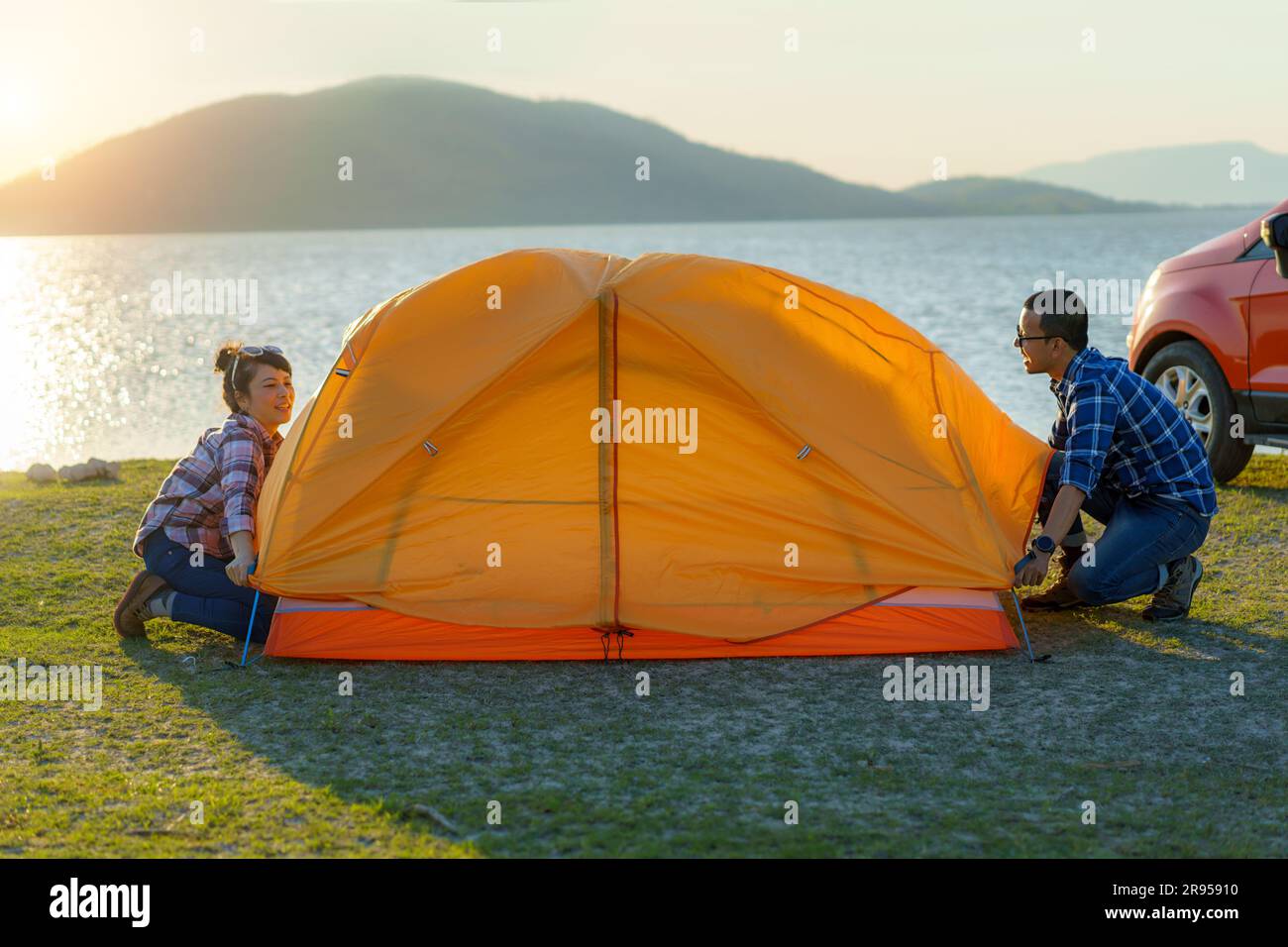 Couple asiatique préparant une tente pour camper dans la pelouse avec le lac en arrière-plan pendant le coucher du soleil Banque D'Images