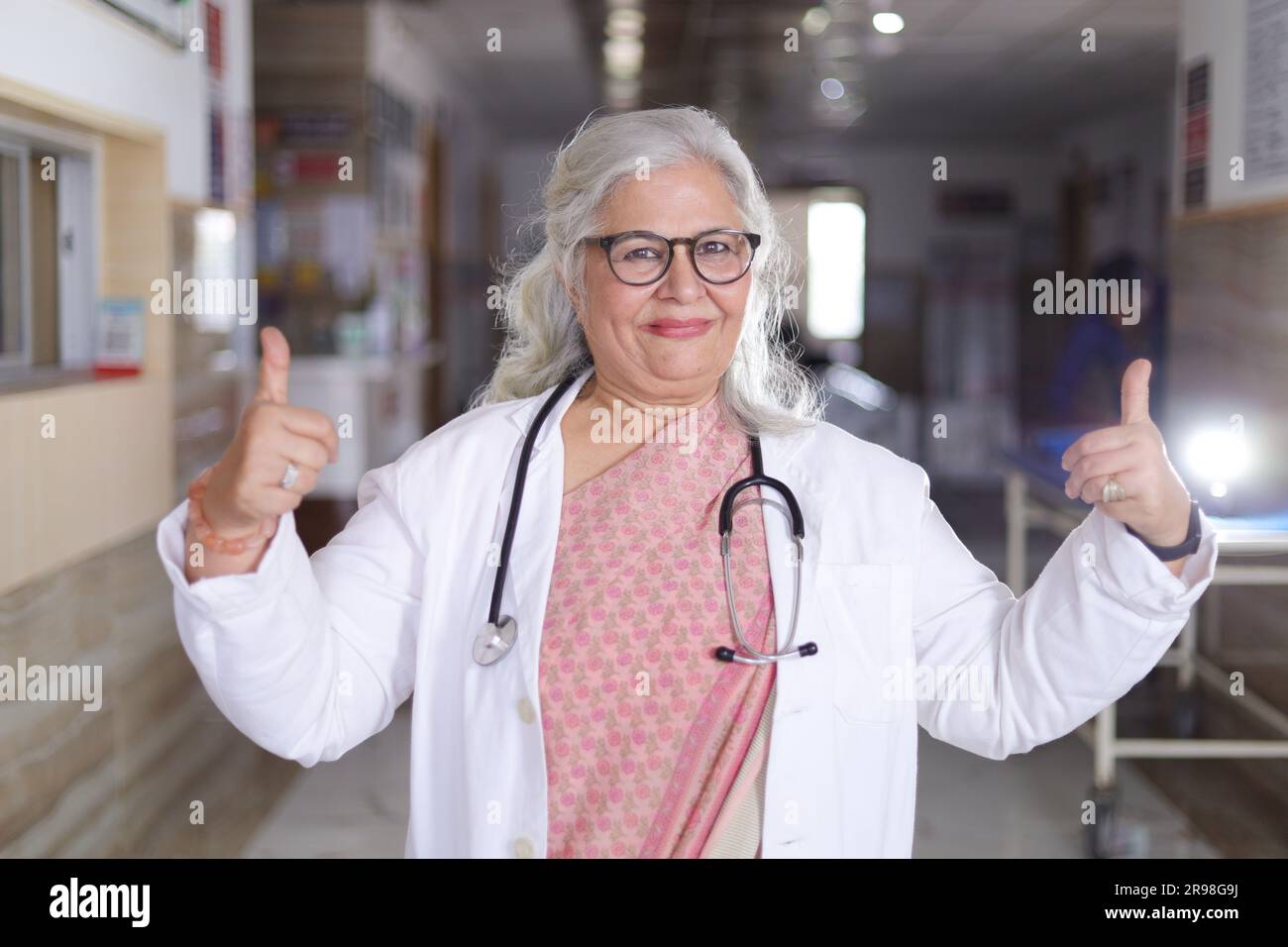 Portrait debout de professionnel médical senior dans le couloir de l'hôpital. Femme indienne docteur décent faisant les pouces vers le haut avec les deux mains. Banque D'Images