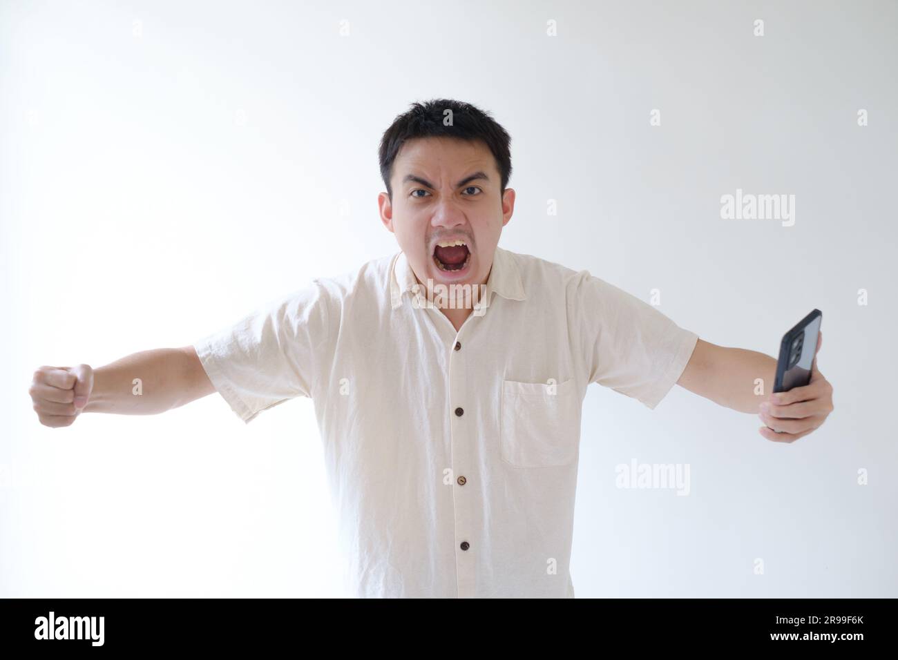 Un jeune asiatique portant une chemise beige et une montre intelligente regarde l'appareil photo avec colère et tient un smartphone. Arrière-plan blanc isolé. Banque D'Images