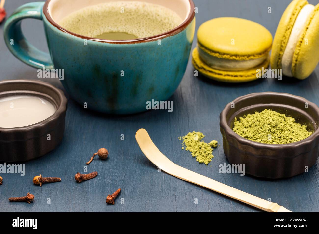 Cuillère de mesure et poudre de matcha verte. Lait dans un bol. Biscuits Macaron et thé matcha dans une tasse. Arrière-plan bleu Banque D'Images