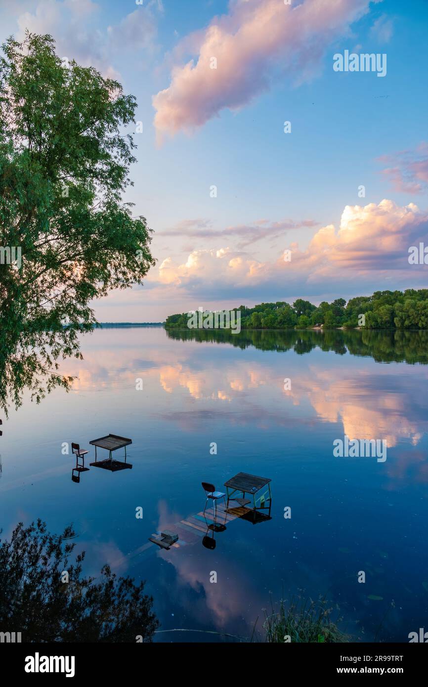 Paysage tranquille au bord de la rivière. Magnifique coucher de soleil sur la rivière avec des points de pêche et des nuages se reflétant dans l'eau. Parc de la ville de Kiev, Ukraine Banque D'Images