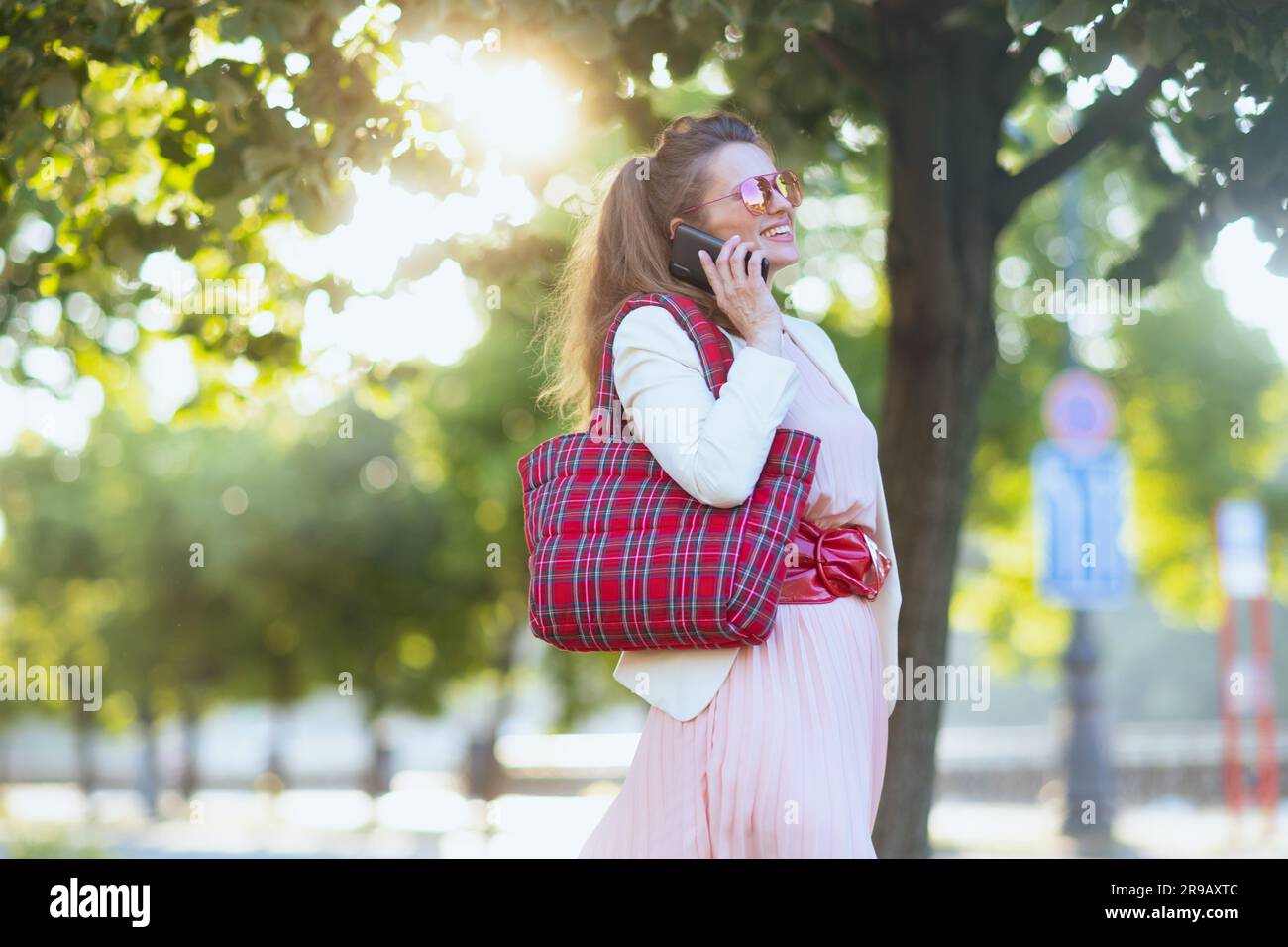 souriante femme de 40 ans en robe rose et veste blanche dans la ville avec sac rouge et lunettes de soleil à l'aide d'un smartphone. Banque D'Images