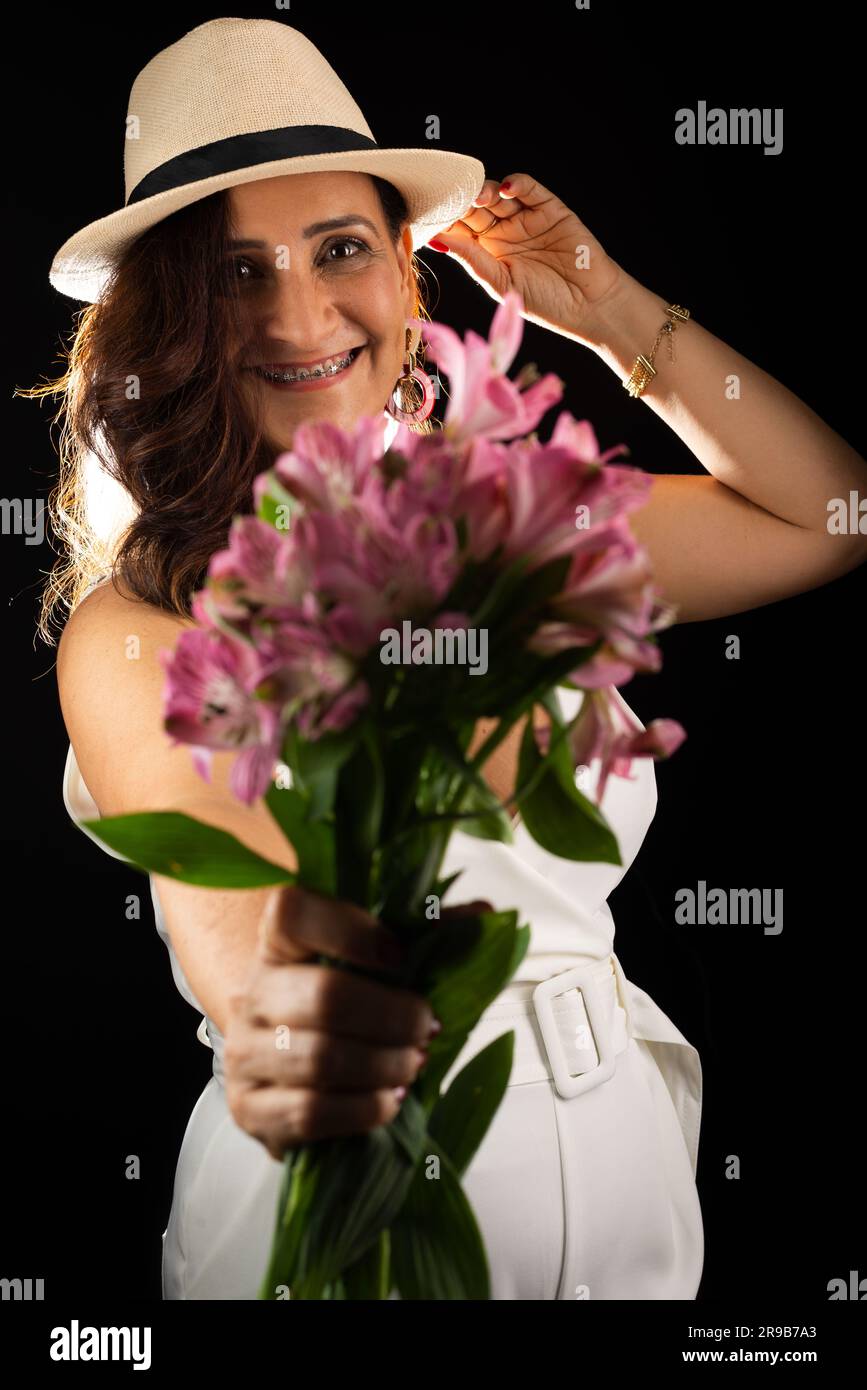 Studio portrait d'une belle femme mûre portant un chapeau tenant un bouquet de fleurs entre les mains. Isolé sur fond noir. Banque D'Images