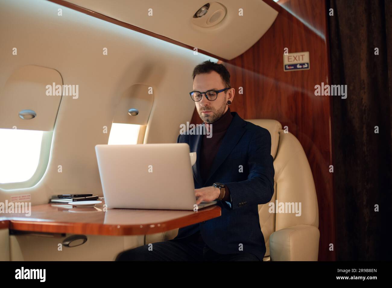 Élégant jeune homme d'affaires PDG dans des lunettes homme assis à l'intérieur d'un avion à jet d'avion privé appréciant le café en travaillant sur son ordinateur portable moderne Banque D'Images