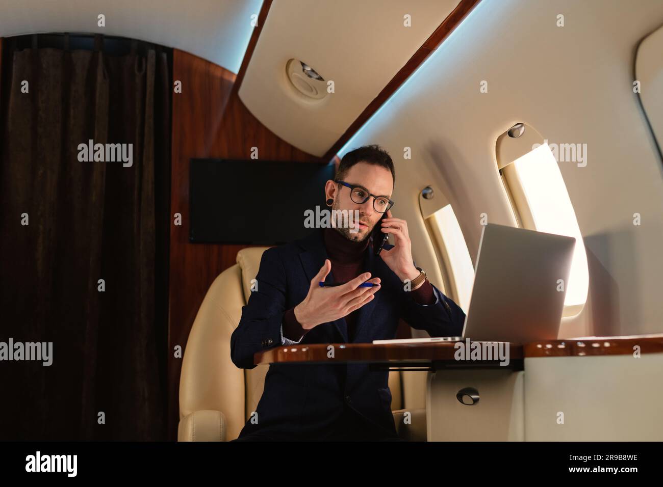 Jeune homme d'affaires élégant dans des lunettes assis à l'intérieur d'un jet d'avion privé parlant sur le téléphone portable et travaillant dans des documents de monnaie papier et ordinateur portable Banque D'Images