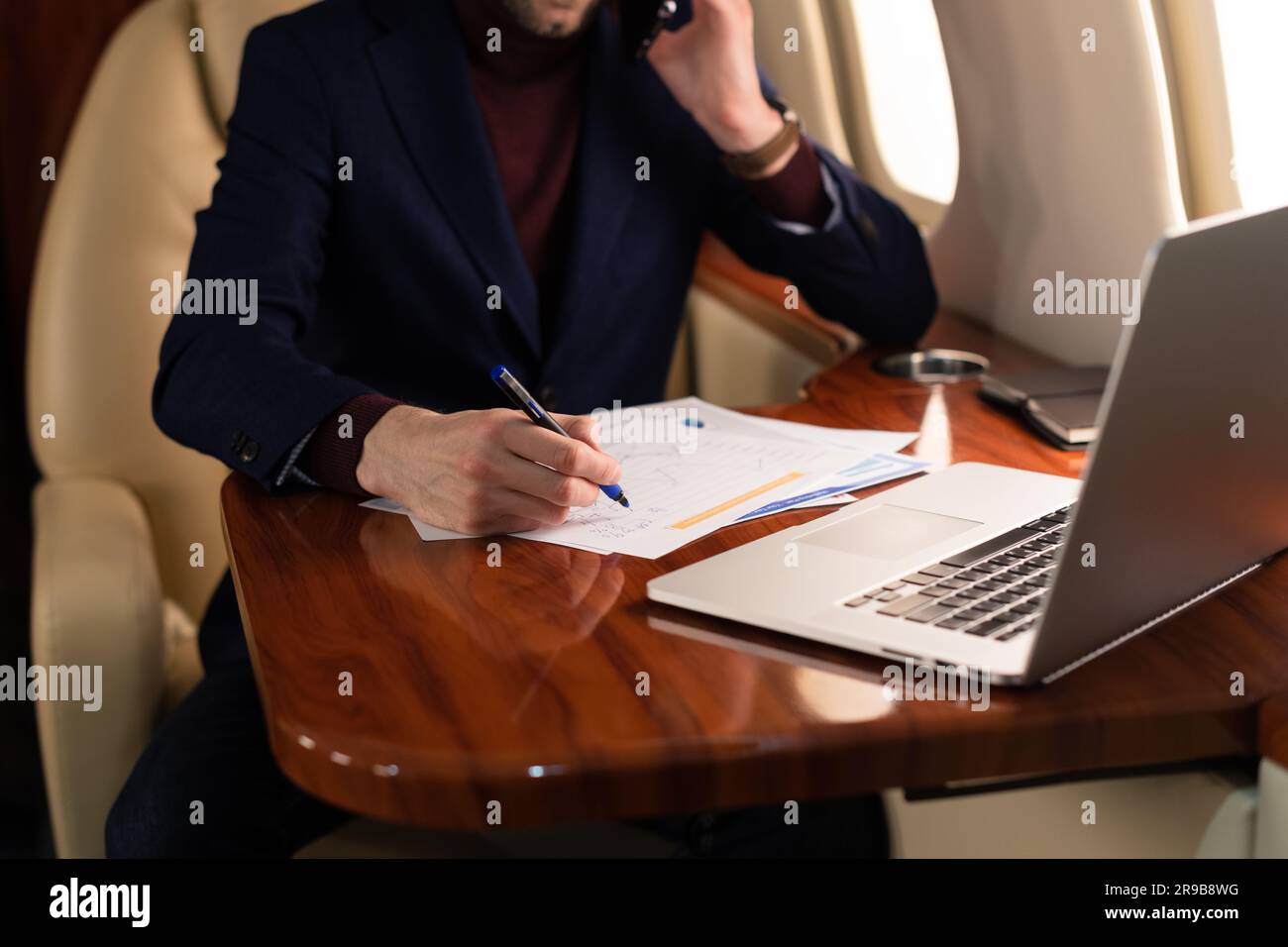 Jeune homme d'affaires élégant PDG dans des lunettes assis à l'intérieur d'un avion à jet privé et travaillant dans des documents de monnaie papier et ordinateur portable pendant le vol Banque D'Images