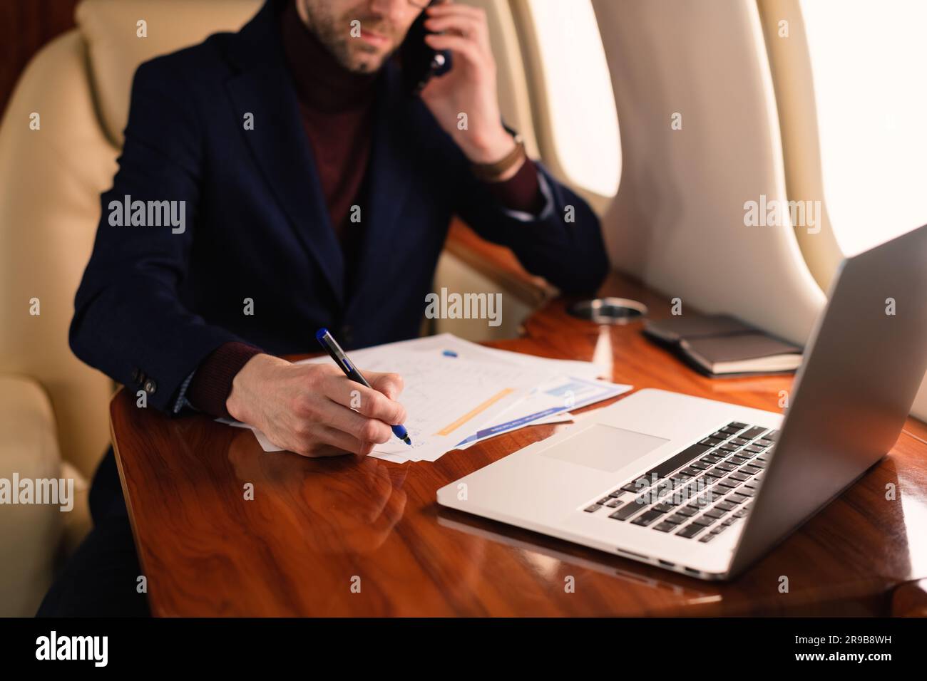 Jeune homme d'affaires élégant PDG dans des lunettes assis à l'intérieur d'un avion à jet privé et travaillant dans des documents de monnaie papier et ordinateur portable pendant le vol Banque D'Images