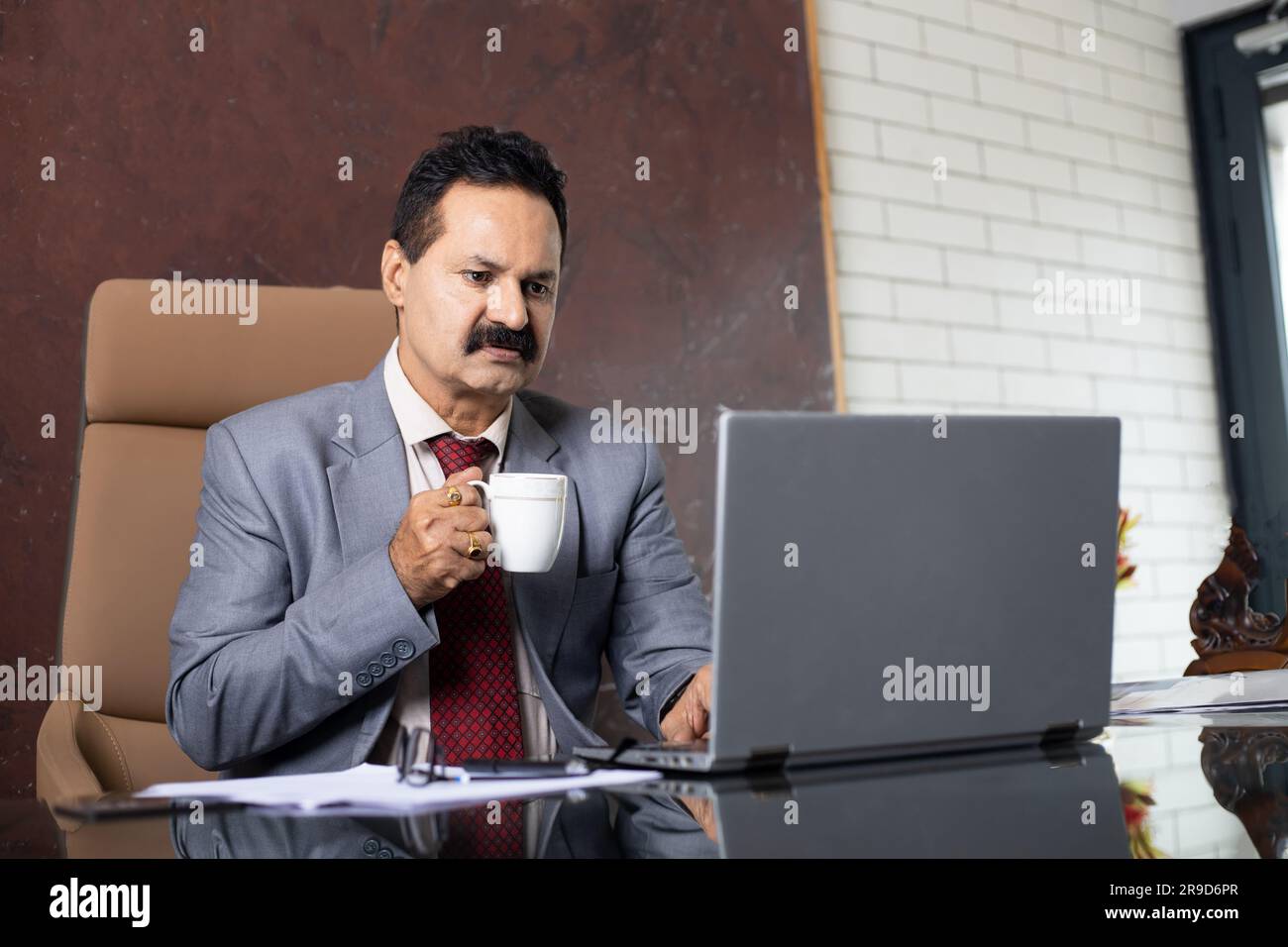 Homme d'affaires indien mûr travaillant sur un ordinateur portable au bureau tout en tenant une tasse de café à la main. Banque D'Images