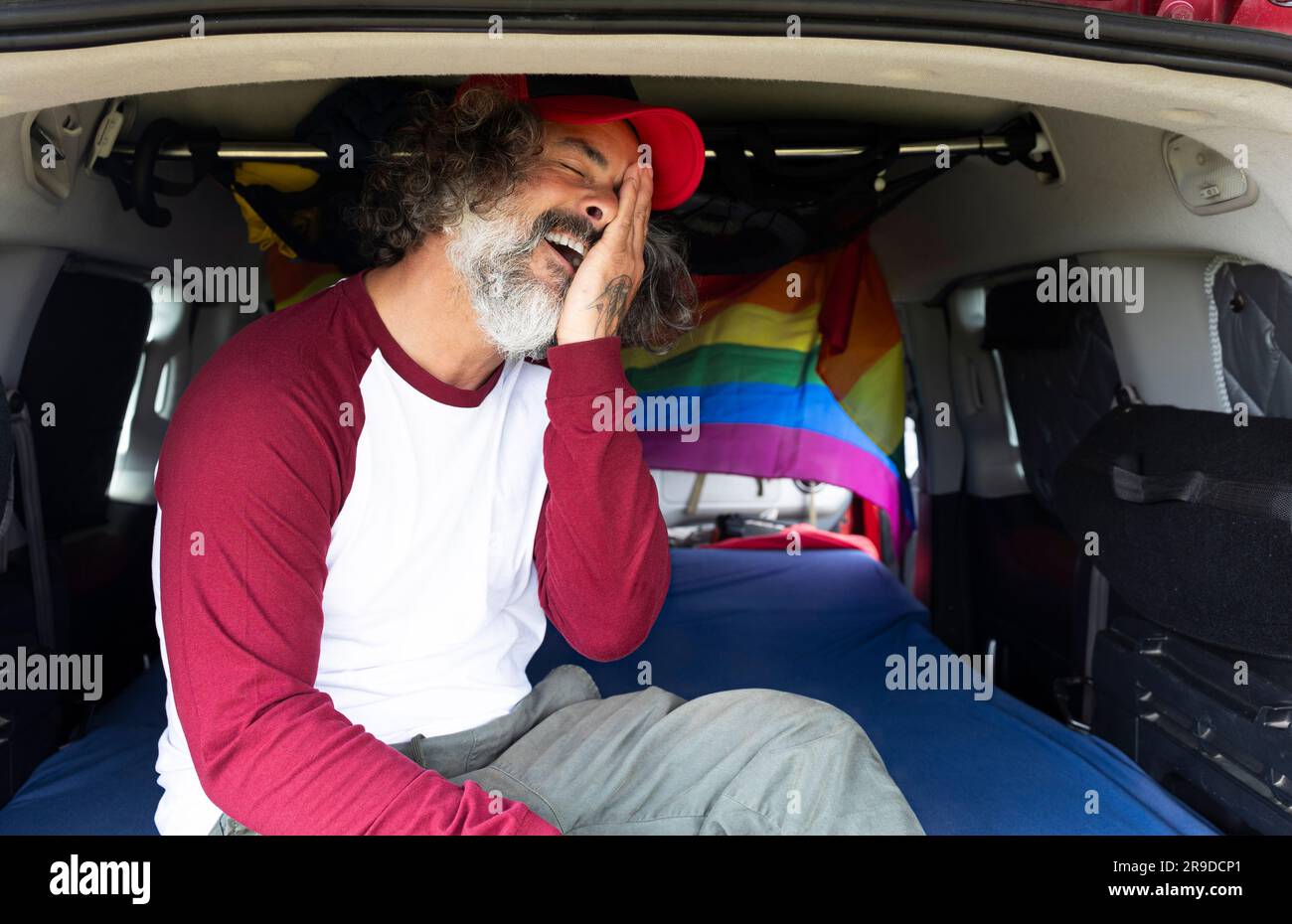 Un homme gay heureux en casquette assis à l'intérieur d'une fourgonnette de camping avec un drapeau de lgtbi Banque D'Images