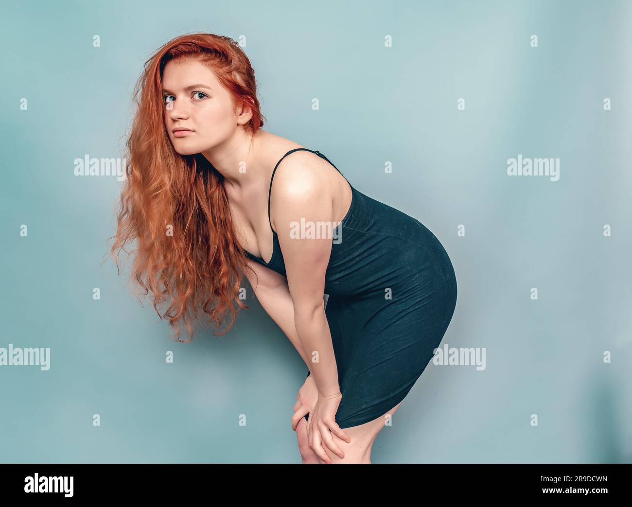 Femme à tête rouge fatiguée posant en studio sur fond gris. Cheveux longs, robe noire Banque D'Images