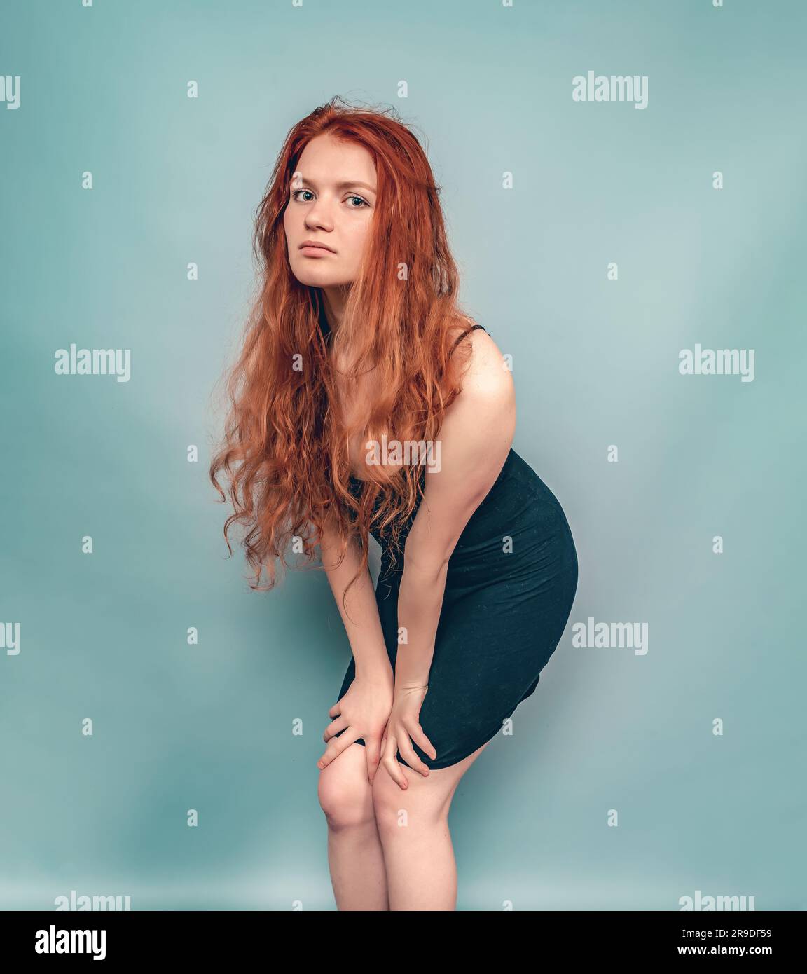 Femme à tête rouge fatiguée posant en studio sur fond gris. Cheveux longs, robe noire Banque D'Images