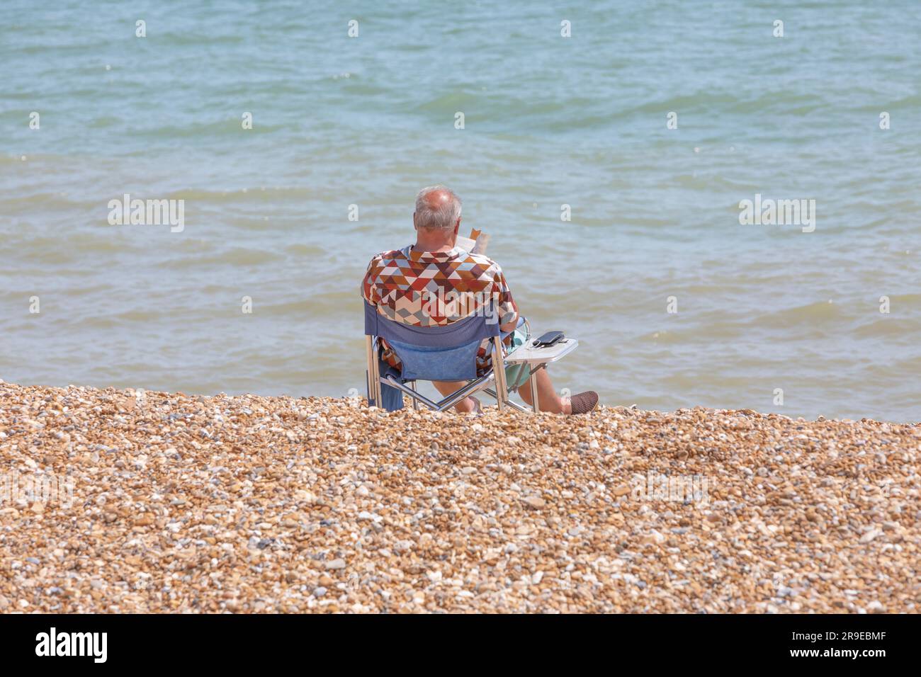 Un homme bronzer et lire un livre assis sur une chaise de plage bleue sur une plage de galets. Banque D'Images