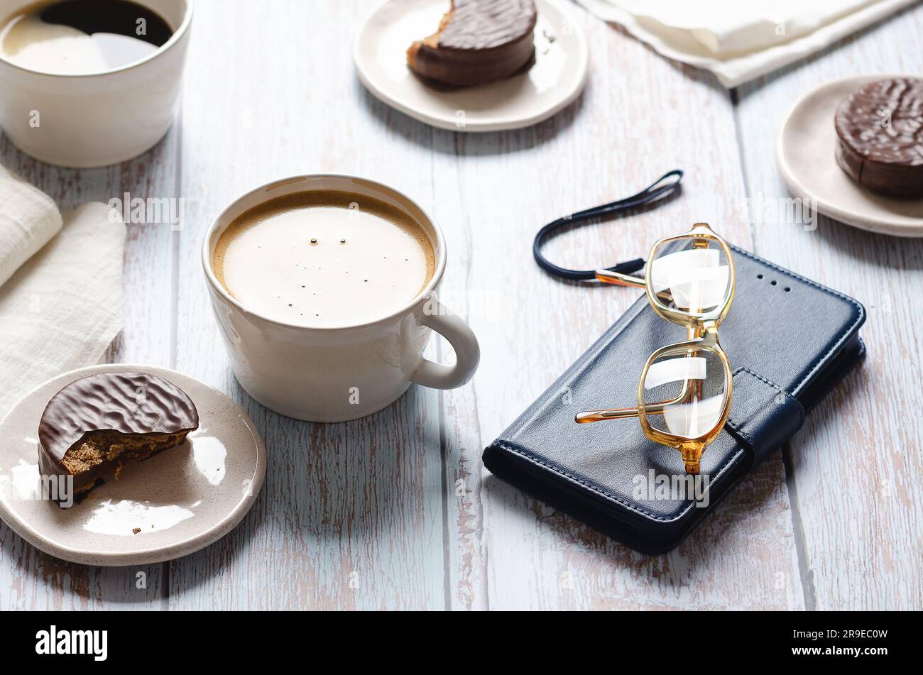 Des tasses de café, des alfajores au chocolat, des verres et un carnet sur une table en bois. Banque D'Images