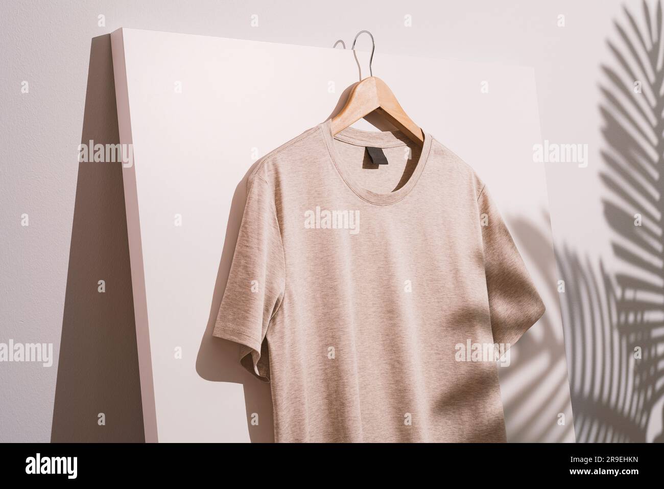Maquette de t-shirt blanc beige, gabarit sur cintre en bois. Ombres des feuilles de palmier en arrière-plan. Banque D'Images