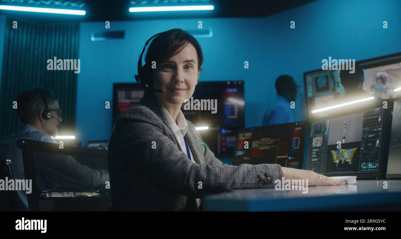 Une employée de contrôle de vol dans un micro-casque se trouve devant l'ordinateur, surveille la mission spatiale dans le centre de contrôle de mission, regarde la caméra. L'équipe regarde la fusée spatiale se lever sur de grands écrans numériques. Portrait. Banque D'Images
