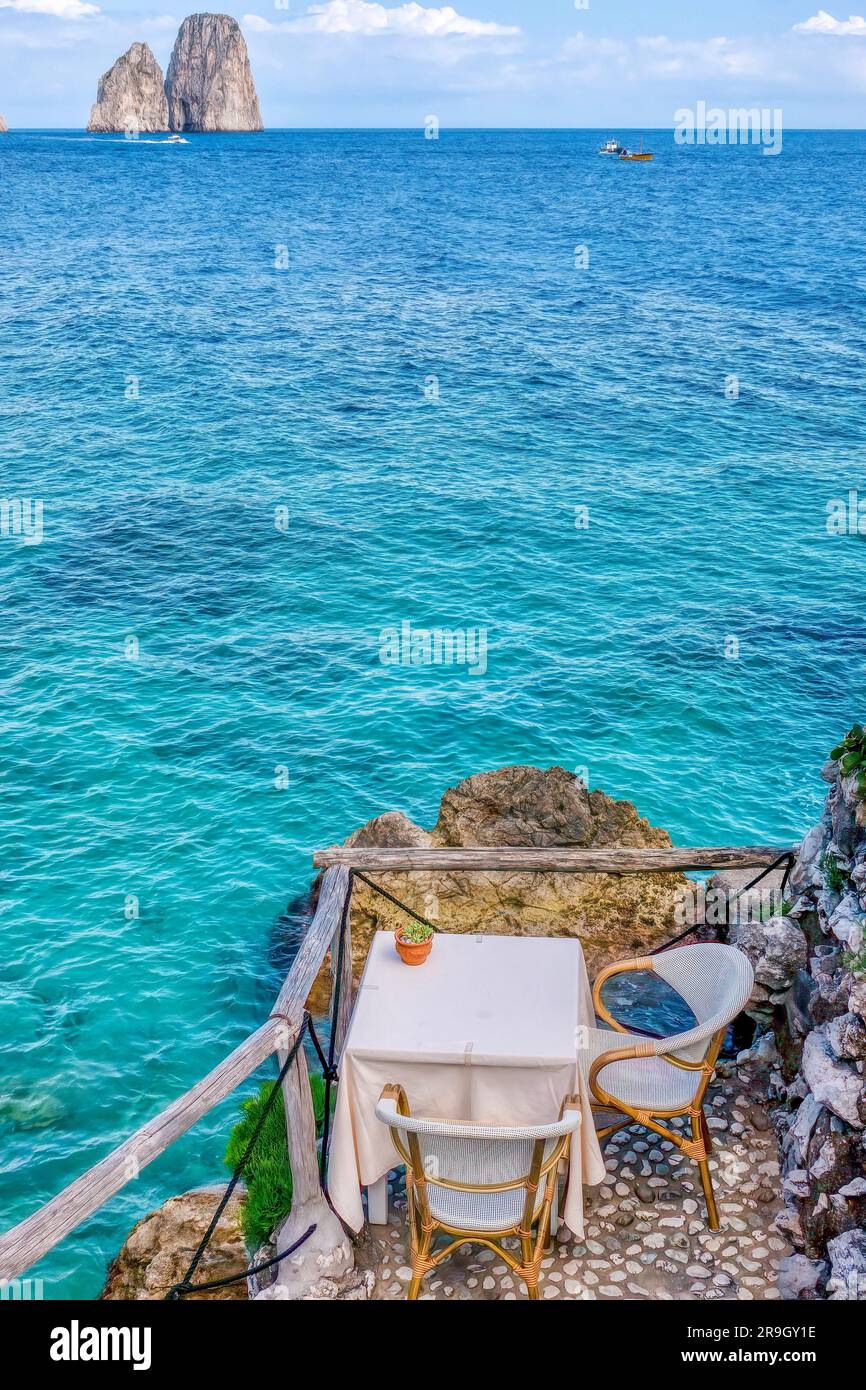 Table pittoresque pour deux personnes sur le front de mer rocheux de l'île italienne de Capri, avec une belle eau bleue et ses deux roches emblématiques de Faraglio en arrière-plan Banque D'Images