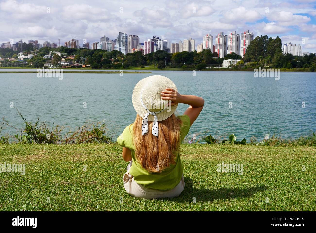 Fille avec chapeau assis sur l'herbe dans Barigui Park appréciant Curitiba paysage urbain, Brésil Banque D'Images