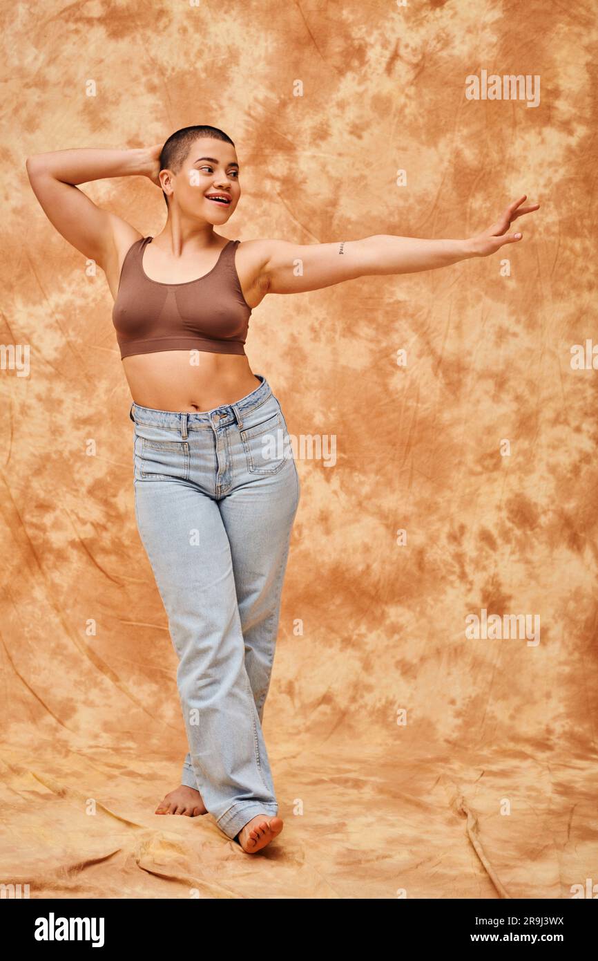 positivité corporelle, look jeans, femme en forme de curvy et heureuse en brassière posant avec une main tendue sur fond beige marbré, tenue décontractée, regarder loin, Banque D'Images