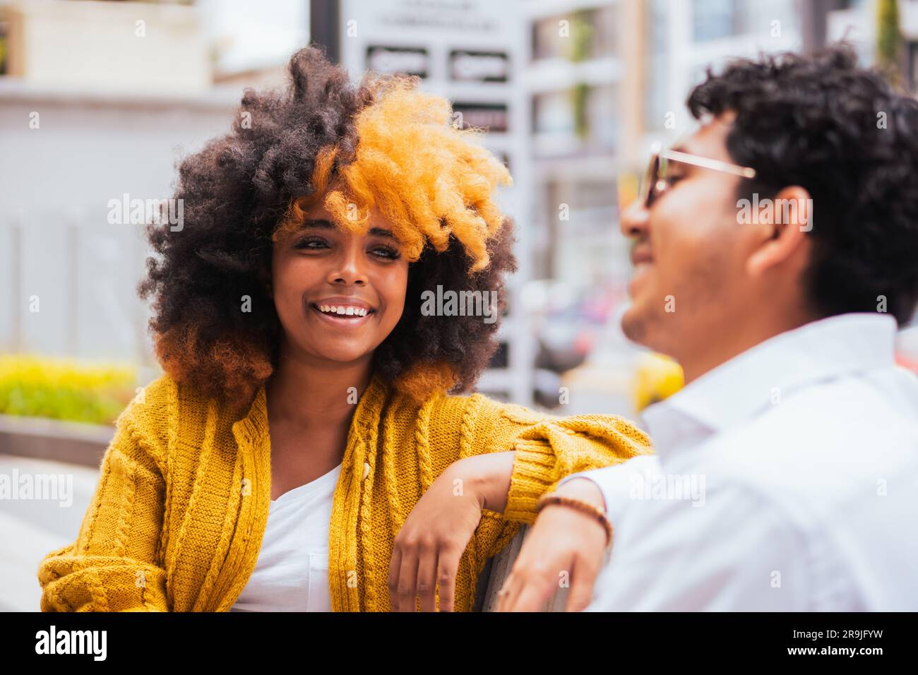 Bonne femme noire avec des cheveux jaunes afro dans des vêtements décontractés souriant et regardant l'ami mâle assis sur un fond flou de la rue de la ville Banque D'Images