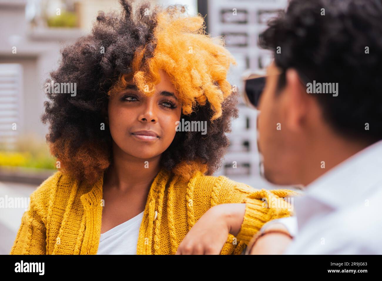 Femme noire aux cheveux jaunes afro dans des vêtements décontractés souriant et regardant un ami mâle assis sur un fond flou de la rue de la ville Banque D'Images