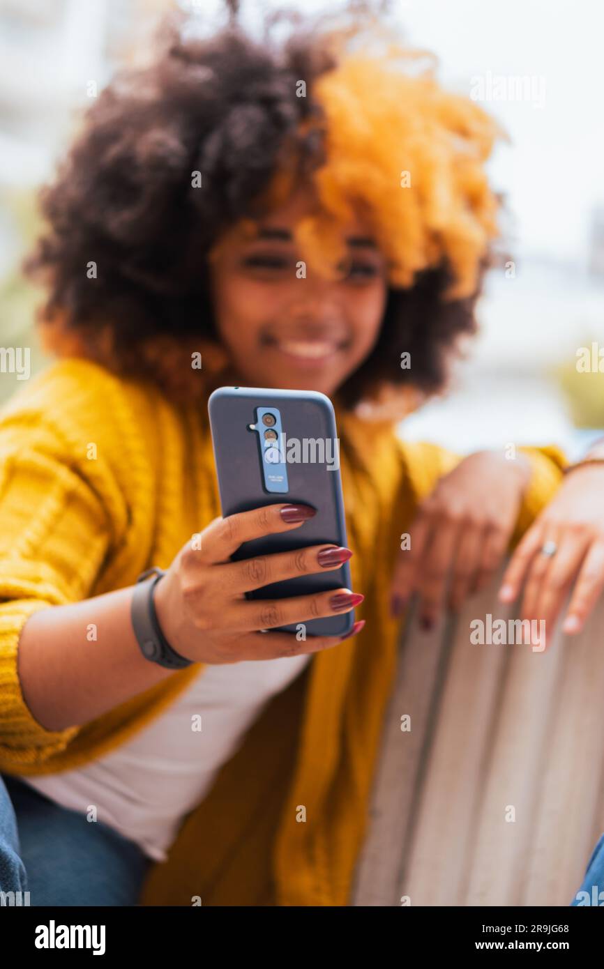 Bonne femme noire avec des cheveux jaunes afro dans des vêtements décontractés souriant et regardant l'écran de smartphone assis sur un fond flou de la rue de ville Banque D'Images