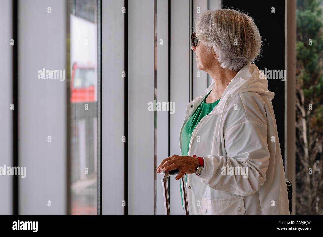 Femme d'âge concentrée avec des cheveux courts gris dans des lunettes de soleil tout en se tenant près de la fenêtre Banque D'Images