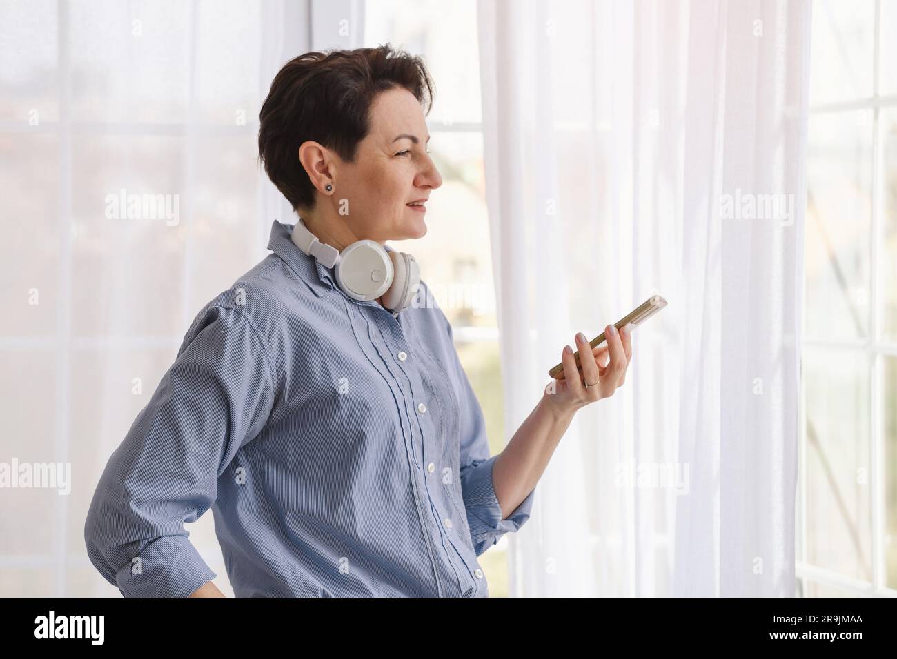 Vue latérale d'une femme d'âge moyen avec des cheveux courts dans une chemise bleue et un casque près de la fenêtre avec un smartphone Banque D'Images