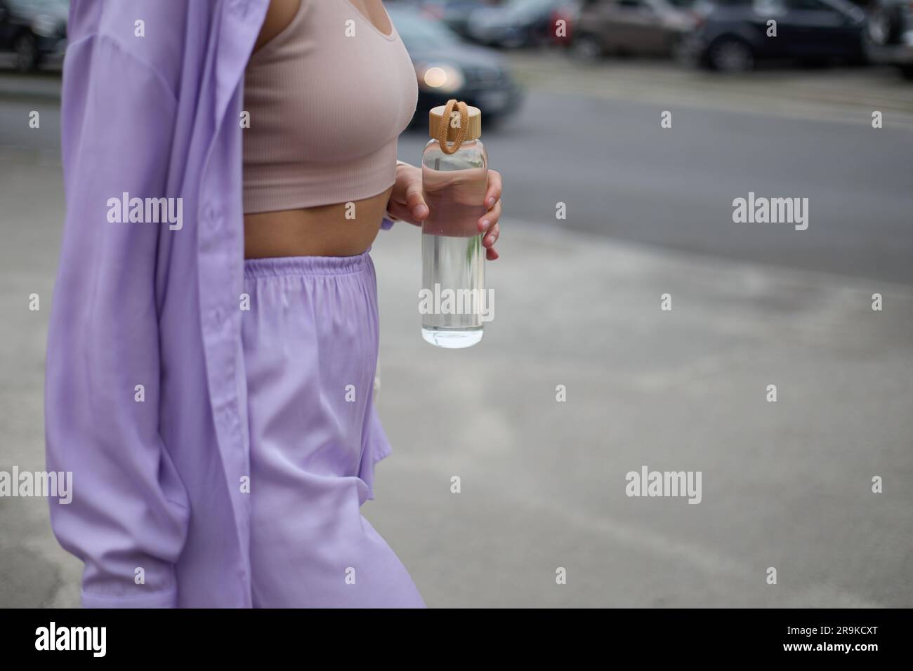 Jeune femme marchant avec une bouteille d'eau en verre à la main. Une femme méconnaissable marche à l'extérieur avec une bouteille réutilisable d'eau minérale à la main. Su Banque D'Images