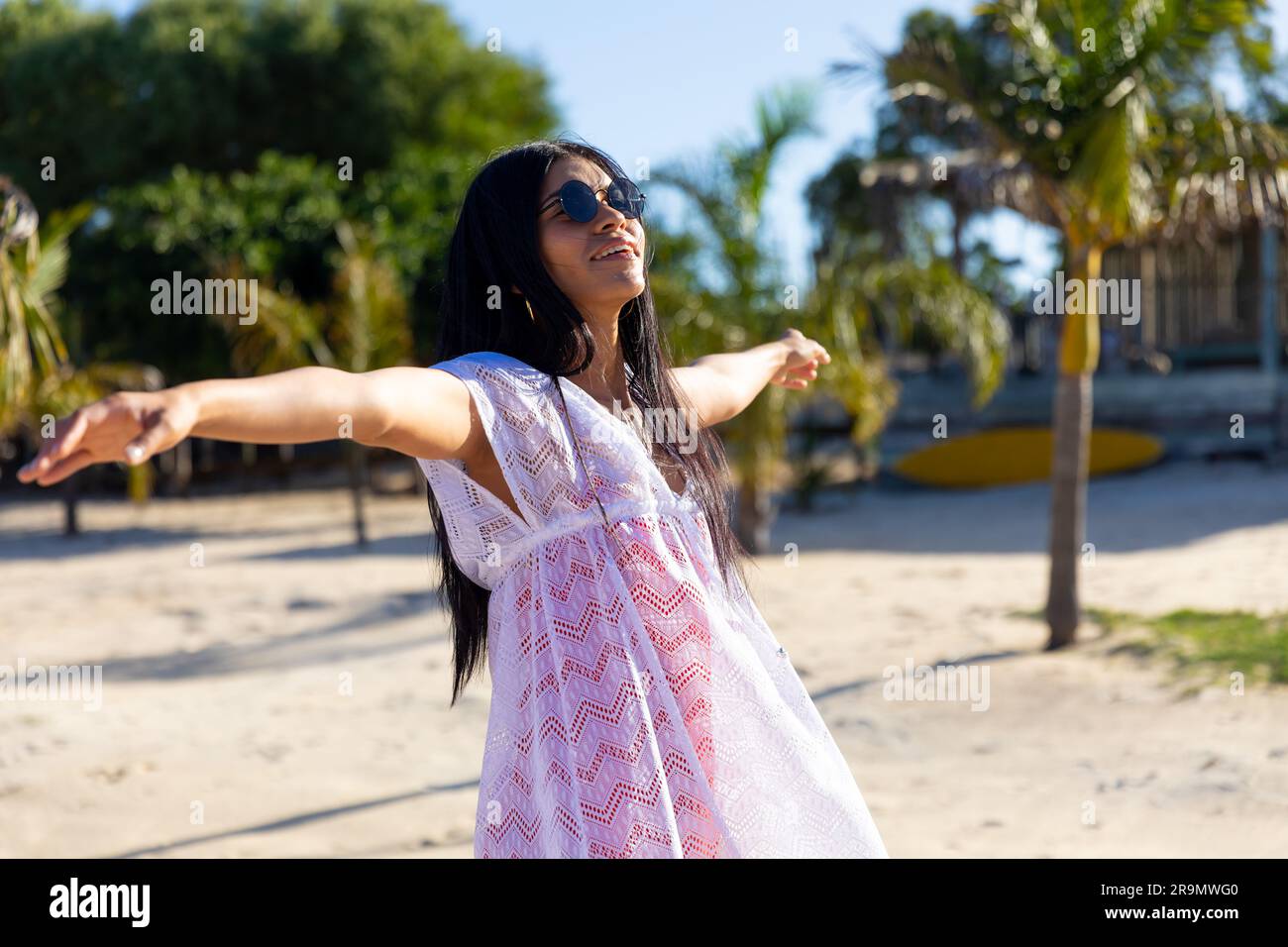 Heureuse femme biraciale dans des lunettes de soleil et robe de plage blanche dansant sur la plage ensoleillée souriante Banque D'Images