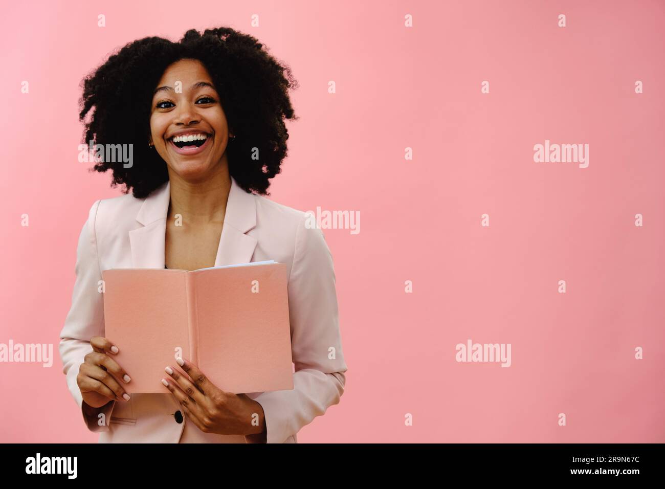 Portrait d'une jeune femme noire tenant un carnet, souriant et riant, sur fond rose de studio Banque D'Images