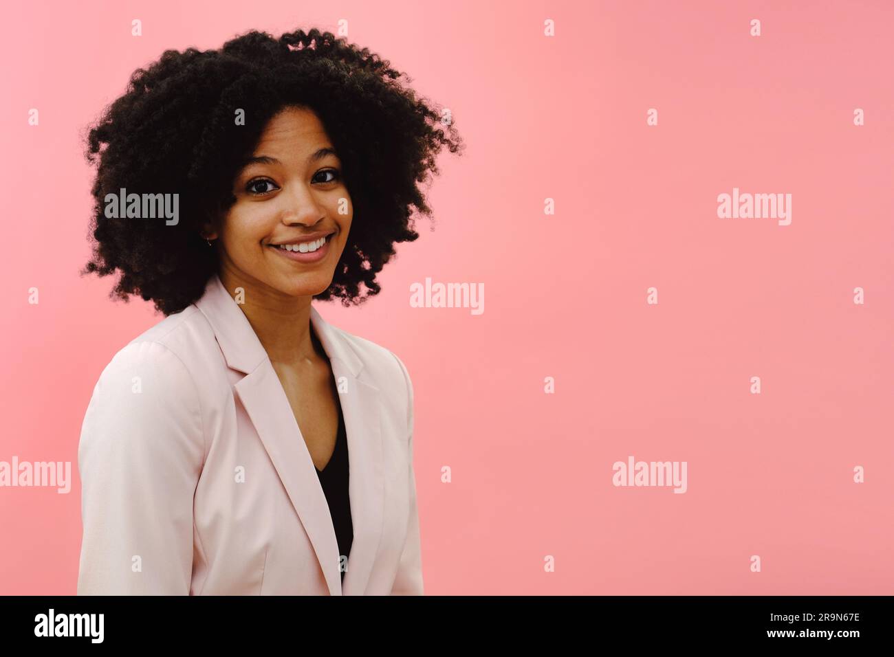 Portrait d'une jeune femme noire portant une veste rose souriant sur fond de studio rose Banque D'Images