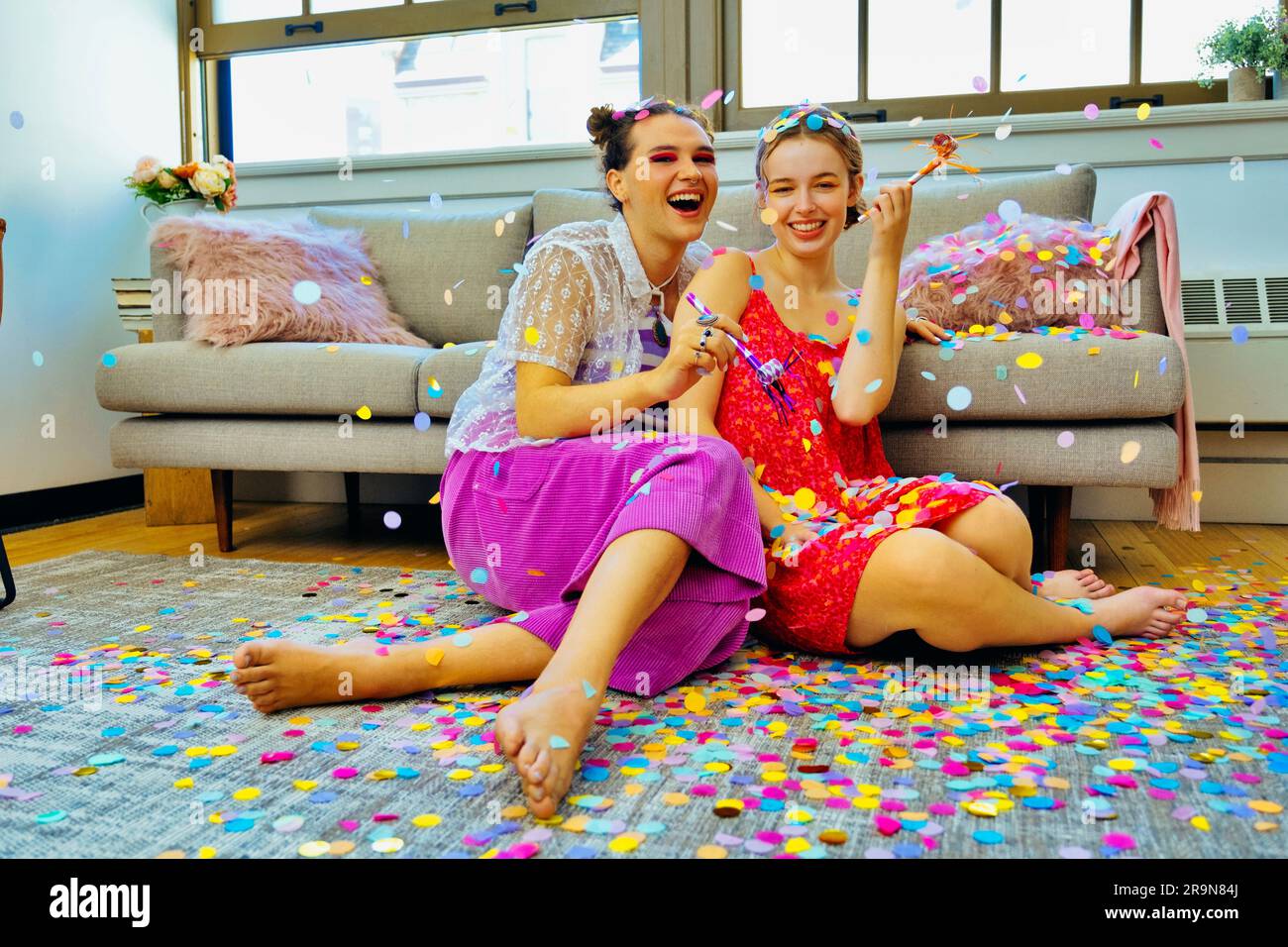 Deux amis qui ont la fête à la maison assis sur le sol tenant des souffleurs de fête avec des confettis tombant autour d'eux. LGBT, non binaire Banque D'Images
