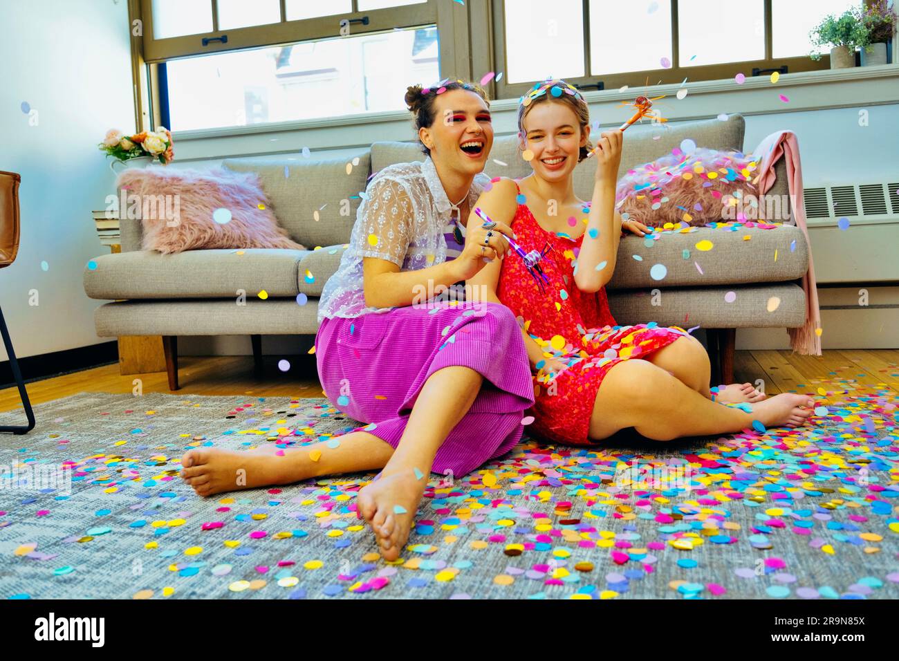 Deux amis qui ont la fête à la maison assis sur le sol tenant des souffleurs de fête avec des confettis tombant autour d'eux. LGBT, non binaire Banque D'Images