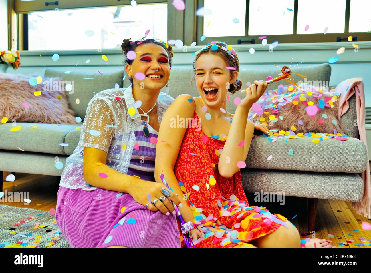 Deux amis qui ont la fête à la maison assis sur le sol tenant des souffleurs de fête avec des confettis tombant autour d'eux Banque D'Images