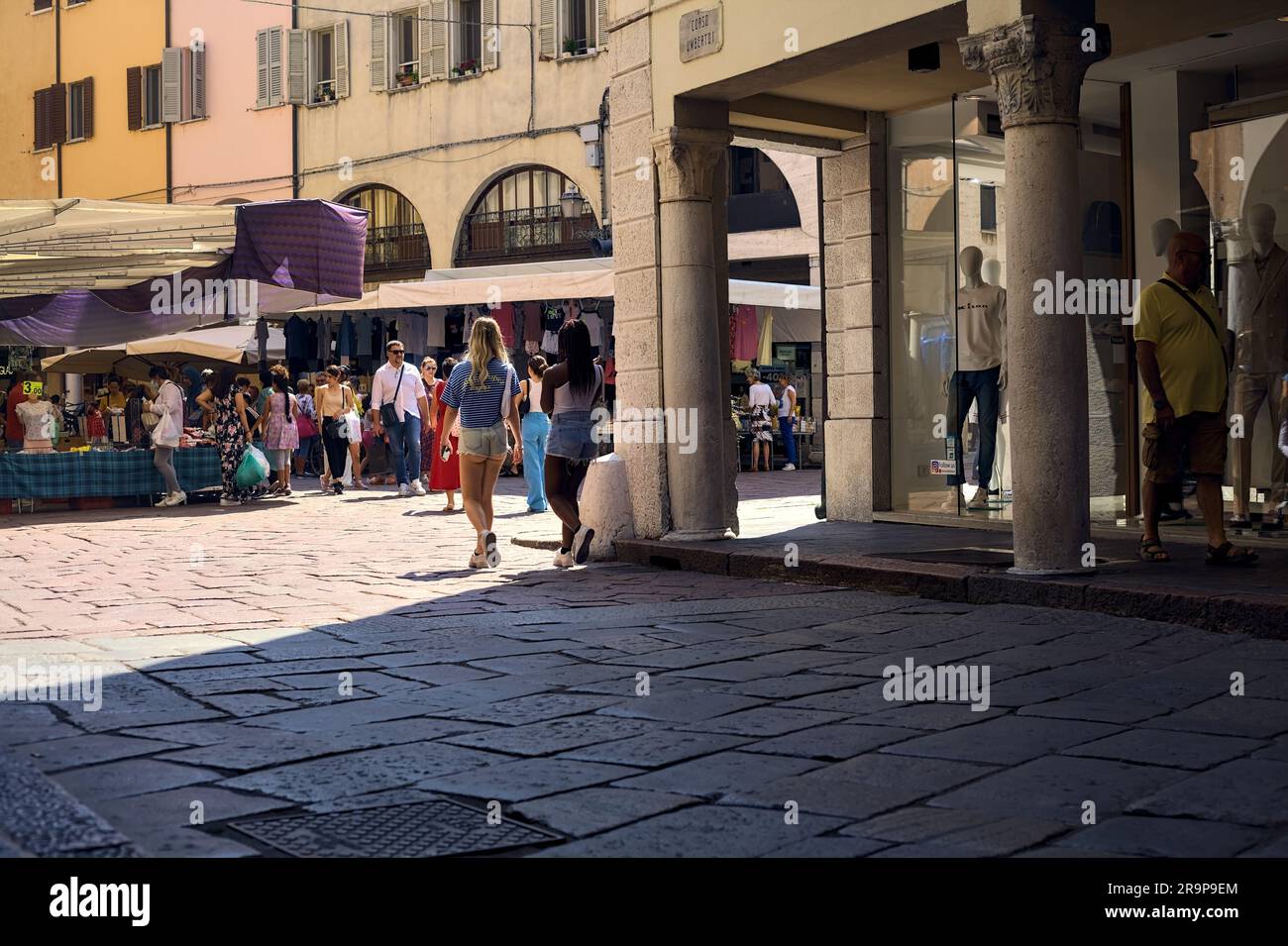 Place avec des stands et des personnes passant par un jour ensoleillé dans une ville italienne vue d'un porche à l'ombre Banque D'Images