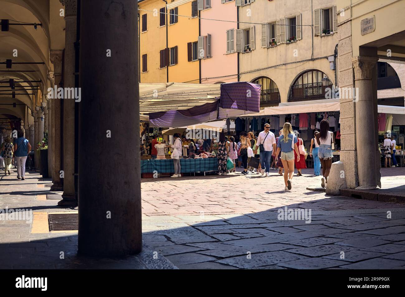 Place avec des stands et des personnes passant par un jour ensoleillé dans une ville italienne vue d'un porche à l'ombre Banque D'Images