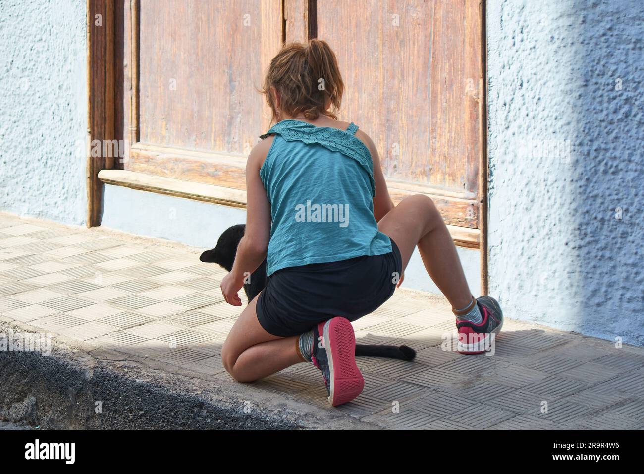 Jeune fille à genoux méconnaissable avec un chat noir devant elle sur une rue ensoleillée. Banque D'Images
