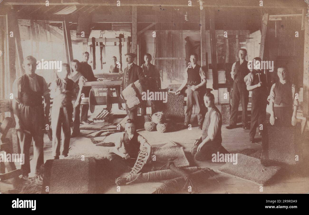 Photographie au début du 20th siècle d'un groupe d'hommes et de garçons dans une usine de fabrication de tapis ou de tapis. Des exemples de tapis fabriqués sont au premier plan et la machine utilisée pour tisser les produits peut être vue vers l'arrière Banque D'Images